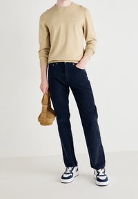 Man in een beige sweatshirt, marineblauwe broek en wit-blauwe sneakers, die een tan kleurige pet in één hand vasthoudt terwijl hij met de andere hand in zijn zak steekt.