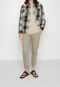 Camicia a quadri nera e crema, t-shirt beige, pantaloni verde oliva e sandali neri con cinturini. La camicia presenta bottoni e tasche.
