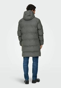Manteau long rembourré en gris foncé avec capuche, présentant une surface texturée et des poignets élastiques, porté sur un jean bleu et des bottes noires.