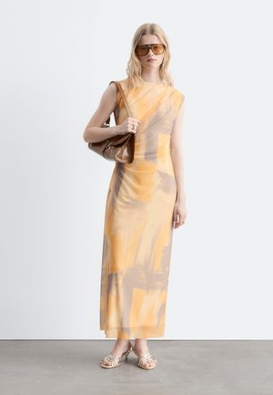 Joven mujer con un vestido maxi de patrón abstracto en amarillo y beige, grandes gafas de sol marrones, sandalias doradas y llevando un bolso de hombro de cuero marrón.