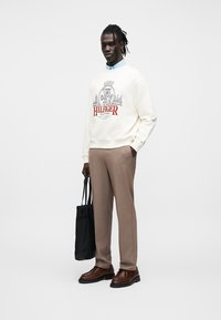 Μπεζ φούτερ με το κόκκινο και γκρι λογότυπο "HILFIGER", μπεζ παντελόνια, καφέ παπούτσια και μαύρη τσάντα ώμου. Κάτω από το φούτερ φαίνεται το γιακά ενός ανοιχτό μπλε πουκάμισου.