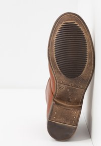Semelle de chaussure en cuir marron avec bande de roulement en caoutchouc usée, marquage taille 37 et couture visible autour des bords, appuyée contre un mur blanc.
