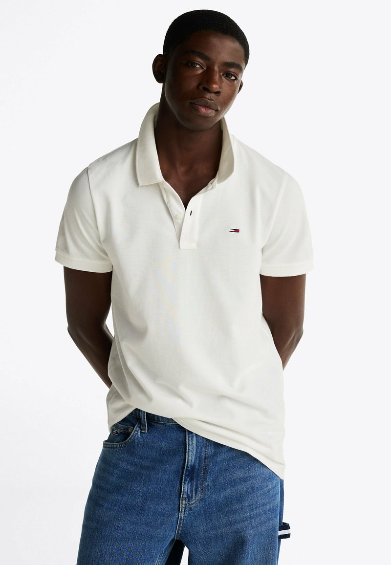 Camisa polo blanca con un collar de dos botones, mangas cortas y un pequeño logo de color en el pecho izquierdo. Combinada con vaqueros de mezclilla azules.