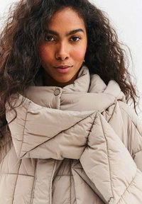 Béžový vatovaný kabát s prošívaným designem, nadměrným límcem a zapínáním na knoflíky, vyrobený z lehkého, texturovaného materiálu.