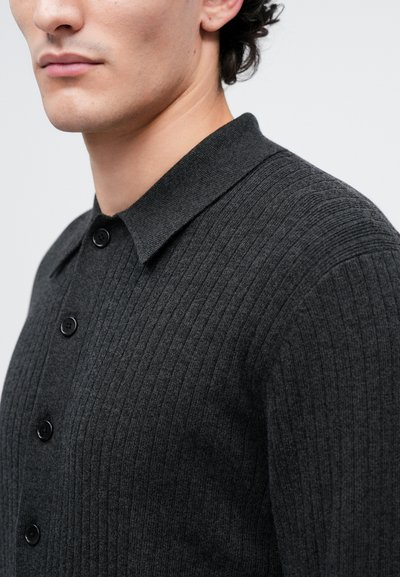 Uomo che indossa un cardigan a coste grigio scuro con collo e bottoni neri, mostrato dalla testa fino a metà tronco.