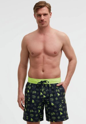 Shirtless man wearing navy blue swim shorts with neon green nautical patterns and waistband, standing with one hand in pocket.
