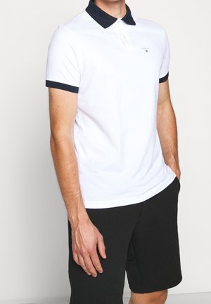 Mężczyzna ubrany w biały polo z czarnym kołnierzem i wykończeniem rękawów, w czarnych szortach, stojący z jedną ręką w kieszeni.