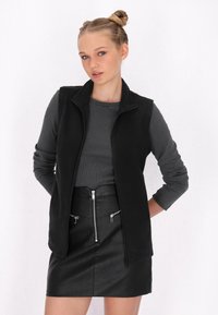 Vest negru fără mâneci, cu fermoar în față, textură moale și guler înalt, purtat cu o cămașă lungă gri închis și o fustă neagră cu fermoar.
