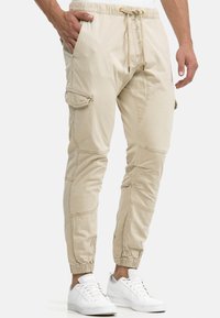 Beige Cargohose mit elastischem Bund, verstellbarem Kordelzug, Seitentaschen und verjüngten Bündchen. Hergestellt aus leichtem Stoff mit weicher Textur.