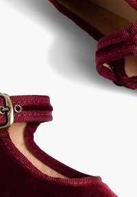 Bordeaux fluwelen schoenen met een ronde neus, enkelbandje en gouden hardware details. Zachte textuur en gladde randen bepalen het ontwerp.