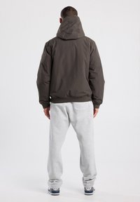 Man facing away wearing a dark hooded jacket, light gray sweatpants, and black and white sneakers on a plain white background.