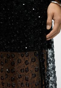 Schwarzes Kleid mit strukturiertem Stoff, verziert mit Pailletten und floralen Mustern. Transparente Überlagerung am unteren Teil zeigt zusätzliche Details.