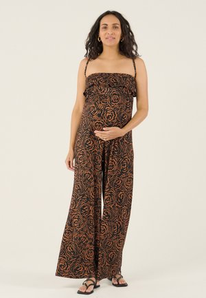 Zwangere vrouw staand, gekleed in een bruin-zwart jumpsuit met bloemenprint zonder bandjes en zwarte sandalen, met één hand op haar buik.