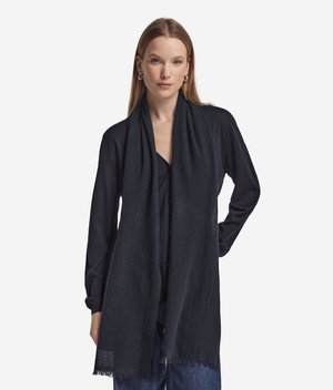 Donna con una lunga sciarpa nera sopra una camicia nera con bottoni e jeans blu, in piedi davanti a uno sfondo bianco semplice.