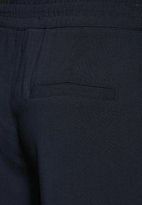 Mörkblått tyg, närbild av byxor med slät struktur, elastisk midja och en enda bakficka med en diskret sydd kontur.