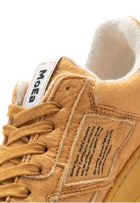 Pruun villast sneaker beezhhi toonides aktsentidega, lapikud paelad, tekstureeritud ülemine osa ja silt, millel on küljes trükitud tekst. Paks tald, ümardatud varvas.
