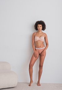 Roza brezšivna modrček in ujemajoče se spodnje hlačke se ponašajo z mehkimi teksturami in minimalnim dizajnom. Model stoji na svetli podlagi z sivo obrobo.