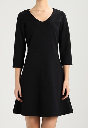 Vrouw draagt een zwarte V-halsjurk met driekwart mouwen en een A-lijn rok, staand tegen een effen lichte achtergrond.