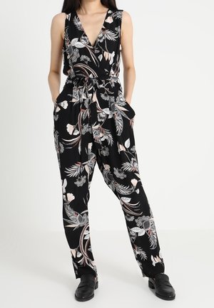 Frau trägt einen schwarzen ärmellosen Blumen-Jumpsuit mit Taillenschnürung, steht mit den Händen in den Taschen und schwarzen Schuhen vor weißem Hintergrund.