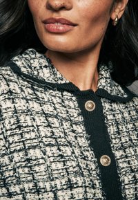 Tweed ülikond, millel on must-valge ruuduline muster, tekstuuriga kangas, kapuuts ja kuldsed nupud. Lähedal vaatamiseks krae ja kaeluse detailid.