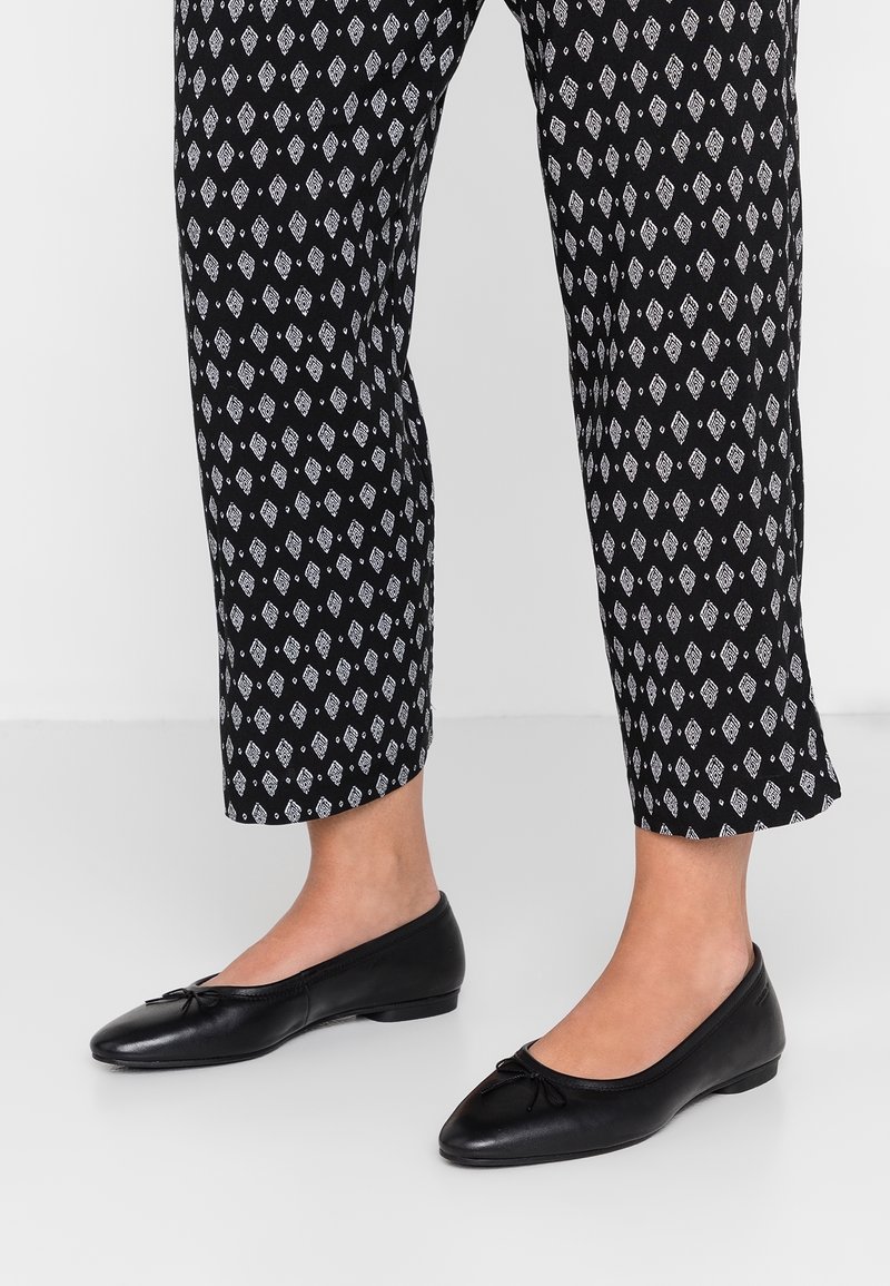 Jambes portant un pantalon court à motifs losanges noir et blanc et des ballerines en cuir noir avec de petits nœuds.
