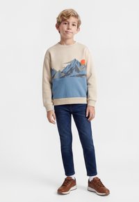 Jongen met een beige en blauw sweatshirt met bergontwerp, donkerblauwe spijkerbroek en bruine sneakers, staand tegen een effen witte achtergrond.
