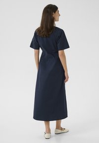 Mornarsko modra obleka do gležnjev s kratkimi rokavi, prilegajočim pasom in gladko teksturo. Nosila se je z svetlimi, koničastimi balerinskimi čevlji.