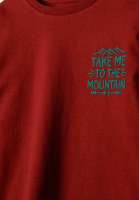 Sarkans krekls ar tirkīza grafiku, kas attēlo kalnu virsotnes un tekstu "Aizved mani uz kalniem" ar dekoratīviem bultu motīviem.