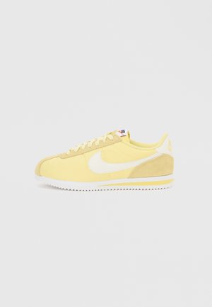 Lage gele en beige Nike-sneaker met wit swoosh-logo en witte zool met gele streep op een effen lichte achtergrond.