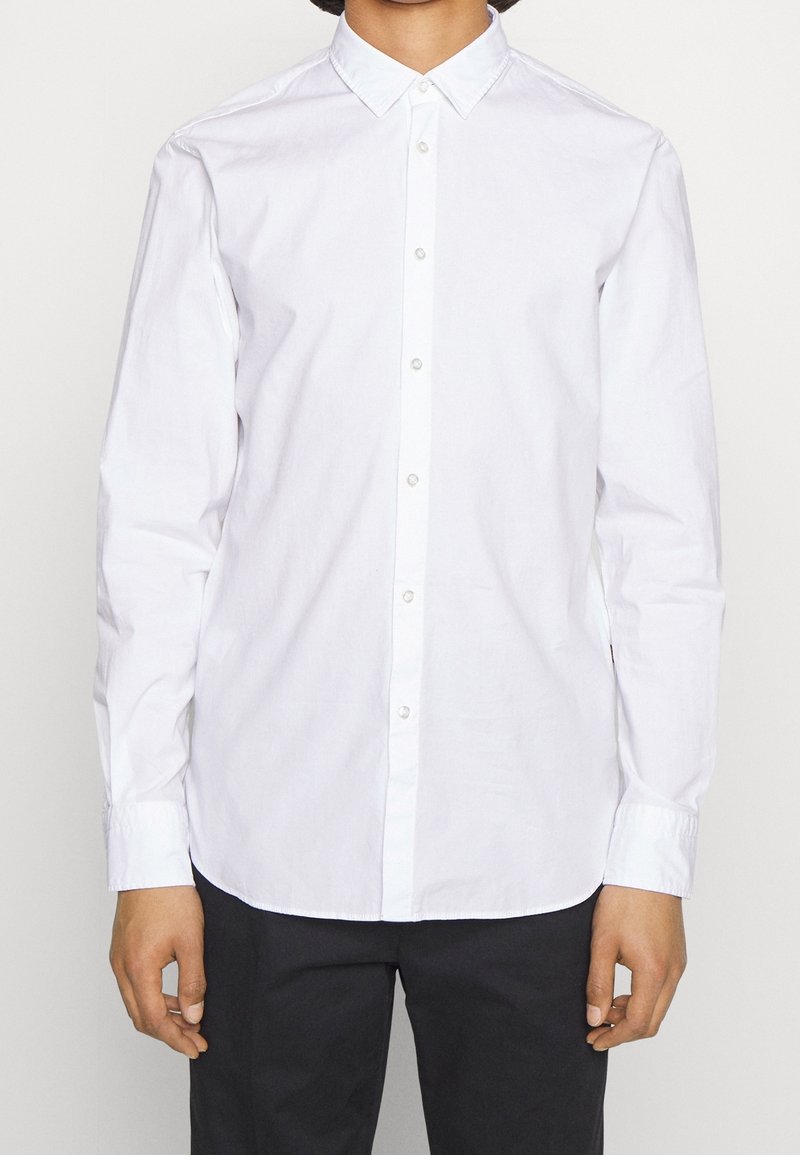 Biała koszula z długim rękawem, kołnierzykiem na guziki, plisą na przodzie z okrągłymi guzikami oraz prostym dołem. Wykonana z gładkiej bawełnianej tkaniny.