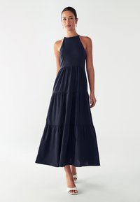 Žena v bezramienkových tmavomodrých maxi šatách s volánmi a bielych sandáloch na otvorenú špičku, stojí na pozadí s jednoduchým svetlým pozadím.