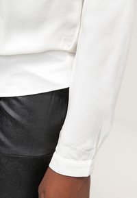 Witte blouse met lange mouwen en een gladde textuur, met knoopmanchetten, gecombineerd met zwarte leren broek, wat de contrasterende kleuren en materialen benadrukt.