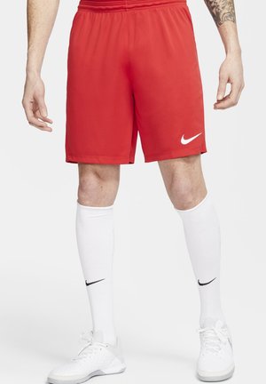 Hombre vistiendo pantalones cortos deportivos Nike rojos, calcetas blancas Nike hasta la rodilla y zapatos deportivos blancos, de pie con las manos relajadas a los lados.