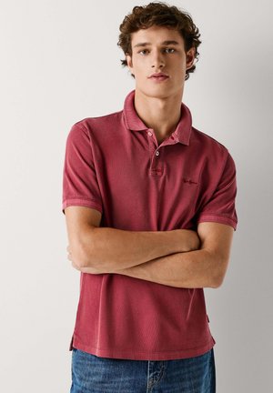 Rotes Poloshirt mit weicher Textur, zwei Knöpfen am Kragen und kleinem Logo. Kombiniert mit blauen Jeans in entspanntem Schnitt.