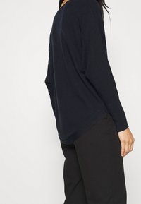 Langärmliger, dunkelblauer Strickpullover in bequemer Passform mit rundem Halsausschnitt und leicht geschwungener Saumlinie, kombiniert mit schwarzen Hosen.