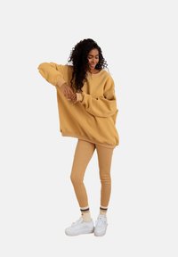 Przesadnie duża żółta bluza z ściągaczami, zestawiona z beżowymi legginsami i białymi trampkami, z paskowanymi skarpetami do kostek.