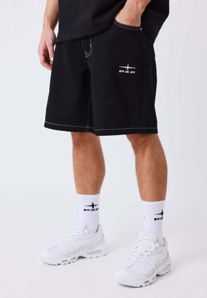 Man draagt zwarte korte broek met wit logo, witte sportsokken met hetzelfde logo en witte sportschoenen, staand met één hand in de zak.