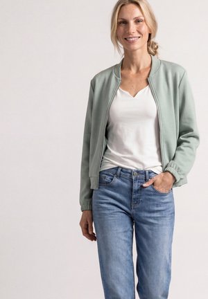 Smilende kvinde med lyst hår i en lav knold iført en lysegrøn lynlåstrøje, hvid skjorte og blå jeans, stående med den ene hånd i lommen.