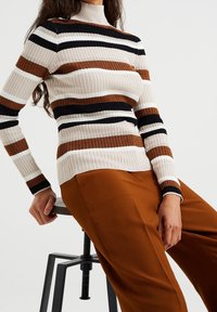 Maglione a costine nei colori crema, nero e marrone, con colletto alto. Abbinato a pantaloni color ruggine, seduto su uno sgabello.