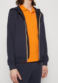 Námořnická modrá mikina na zip se žlutými akcenty, nošená přes oranžové polo tričko. Zobrazená s bočními kapsami a žebrovanými lemy rukávů. Hladká struktura látky.