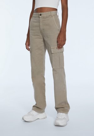 Person wearing beige cargo pants with side pockets and white sneakers, standing against a plain light background.