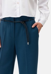 Teal op maat gemaakte broeken met een zwarte koordriem, voorzien van zijzakken en een geplooid ontwerp aan de voorkant. Gemaakt in combinatie met een witte cropped shirt.