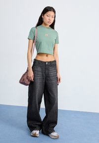 Cropped green cotton t-shirt with front logo, paired with loose black denim cargo pants. Accented with white sneakers and a pink shoulder bag.