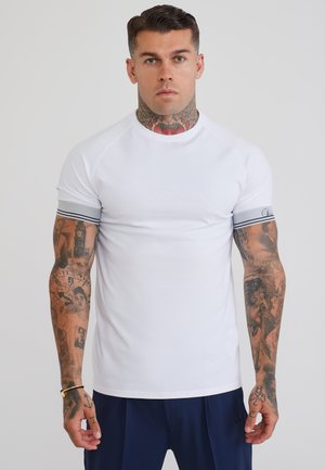 Man met tatoeages op nek en armen, met een wit kortemouwenoverhemd met gestreepte manchetten en marineblauwe broek, staande tegen een effen achtergrond.