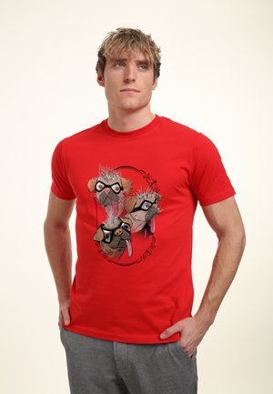 Jeune homme en t-shirt rouge avec un dessin de trois carlins de dessin animé avec lunettes et langue tirée, debout les mains dans les poches d’un pantalon gris.