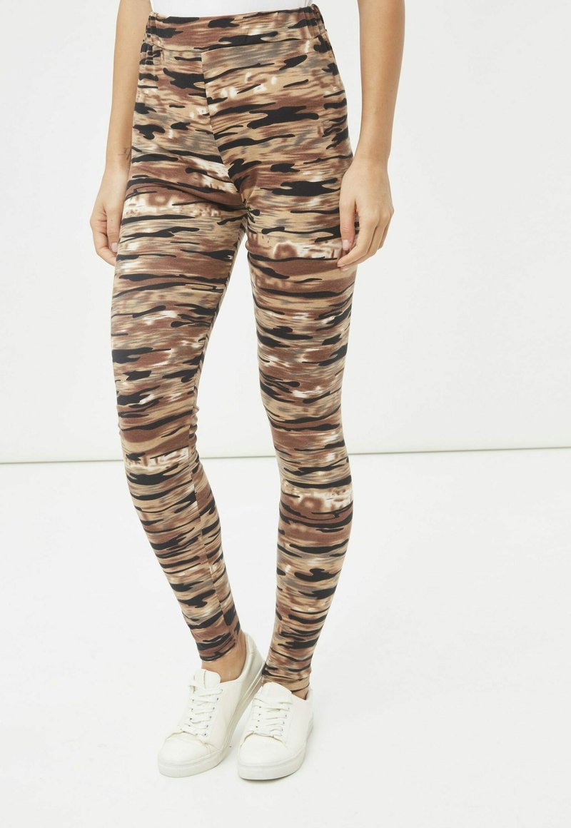 Mujer que lleva leggings de rayas abstractas en marrón y negro con zapatillas blancas, de pie frente a un fondo blanco liso.