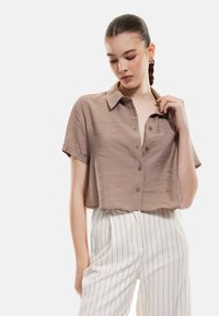 Lyhyt, cropped-sedekshirt vaaleanruskeassa värissä, valmistettu teksturoidusta kankaasta, jossa on napitettava etuosa ja terävä kaulus.