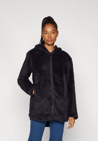 Schwarzer, flauschiger, langer Hoodie mit Reißverschluss vorne, Kapuze und zwei vorderen Taschen. Weicher Stoff mit lockerer Passform und abgerundetem Saum.