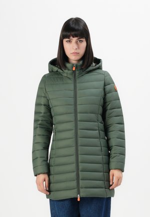 Jonge vrouw in een lange, gewatteerde groene jas met capuchon en oranje ritssluitingen, staande tegen een effen witte achtergrond.