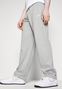 En person i ljusgrå Nike sweatpants och vita Nike sneakers med svarta sulor, stående på ett vitt golv mot en enfärgad bakgrund.